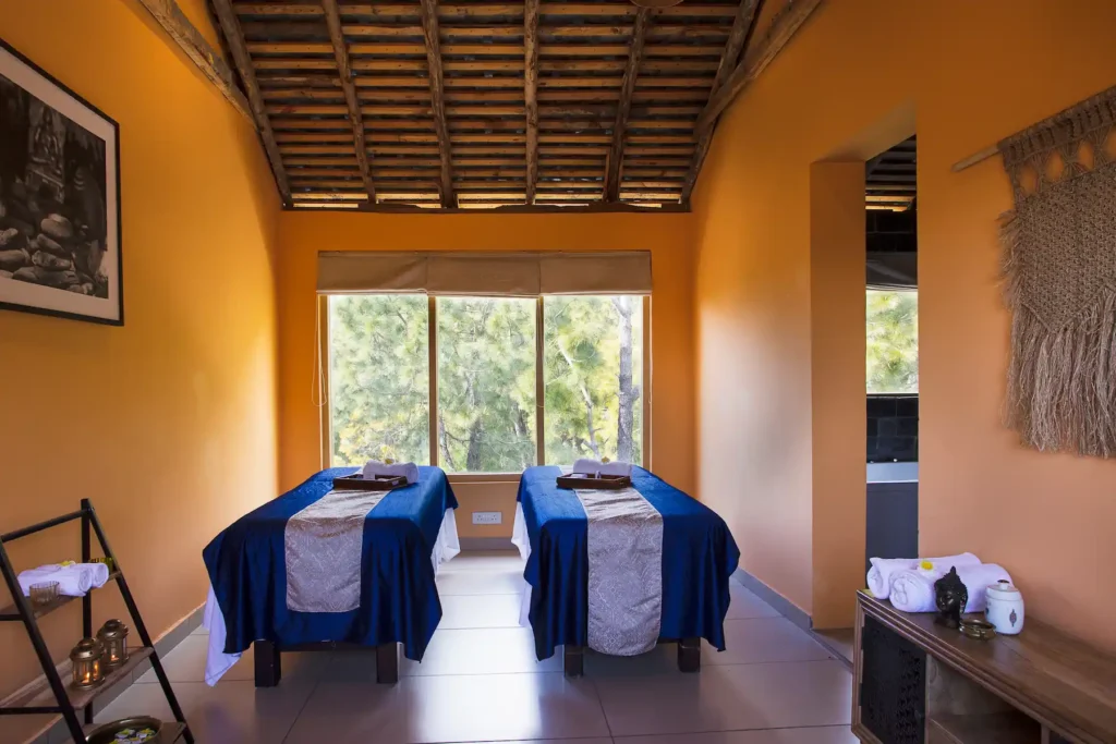 A serene spa treatment room with two massage beds draped in blue linens, warm orange walls, and a large window overlooking green trees.